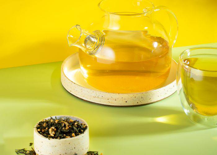 A glass tea pot of genmaicha tea, a golden yellow color that's complemented by the yellow background and green surface. In the foreground, genmaicha tea leaves in a bowl.